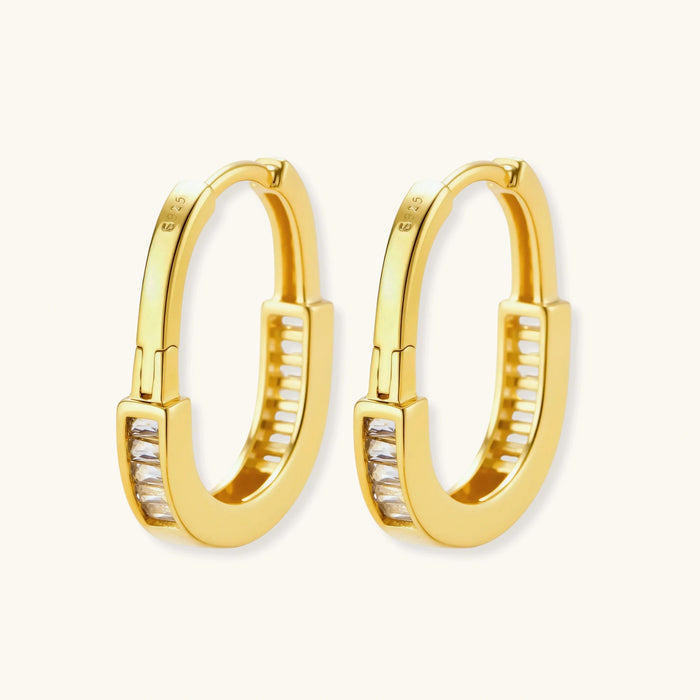 Close-up of gold-plated sterling silver huggie hoop earrings featuring rectangular cubic zirconia stones on neutral background.